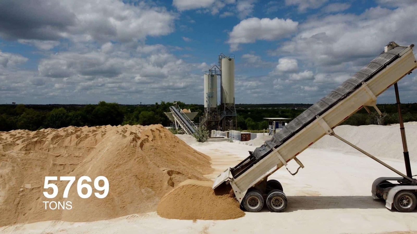 A dump trailer unloads sand onto a stockpile at a materials yard, with two silos and a conveyor in the background and an illustrative figure, 5769 tons, overlaid on the pile.
