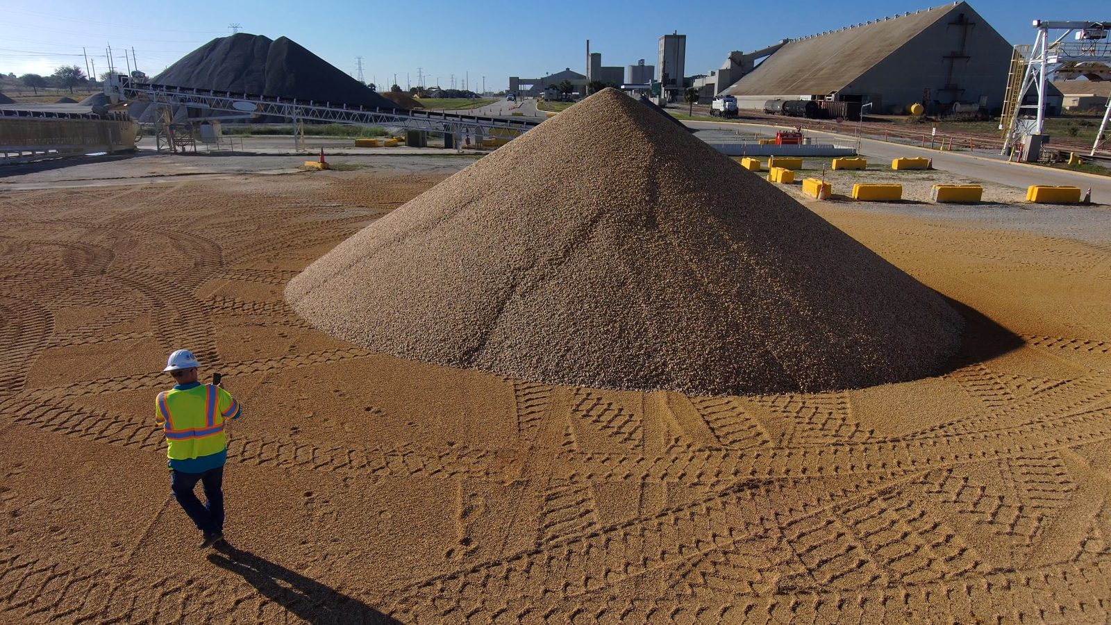 A worker in a hi-vis vest and hard hat walks across the swept pad of a materials yard toward a large conical aggregate pile, with conveyors, silos and covered storage behind.