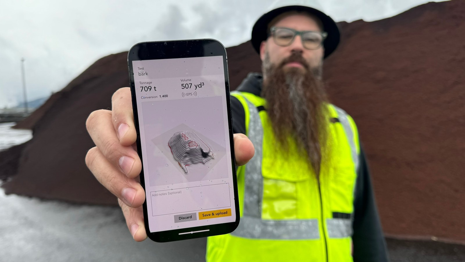 A worker in a high-visibility vest holds up a phone showing a completed measurement of 709 tons and 507 cubic yards, standing in front of the bark pile it measured.