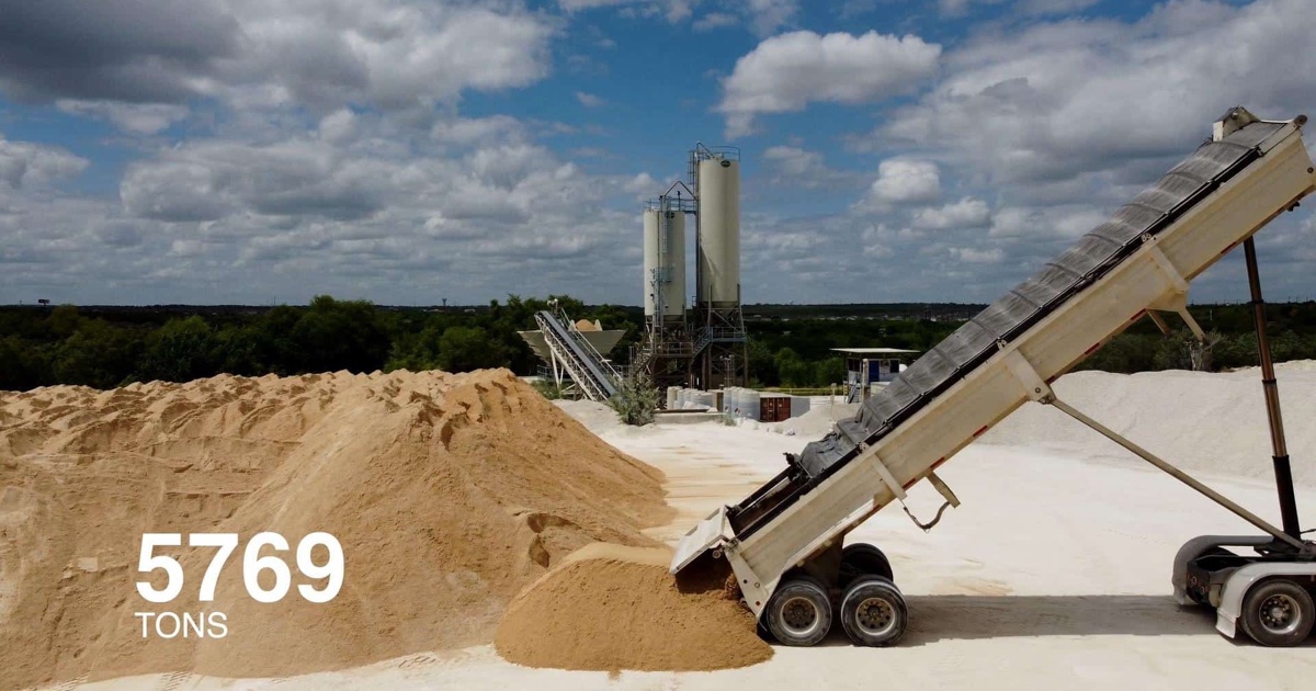 A dump trailer unloads sand onto a stockpile at a materials yard, with two silos and a conveyor in the background and an illustrative figure, 5769 tons, overlaid on the pile.