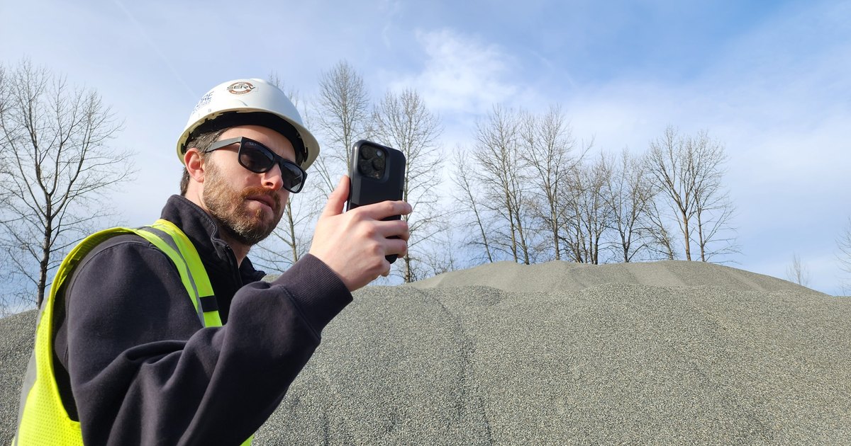A worker in a hard hat and high-visibility vest measures a stockpile of crushed aggregate with an iPhone.
