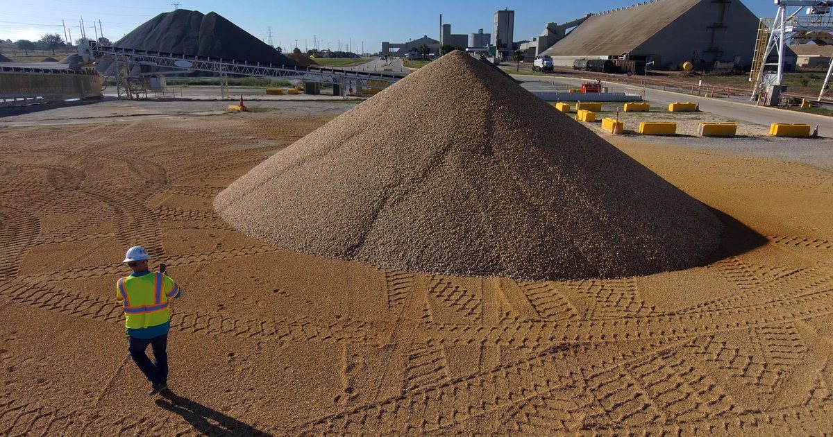 A worker in a hi-vis vest and hard hat walks across the swept pad of a materials yard toward a large conical aggregate pile, with conveyors, silos and covered storage behind.