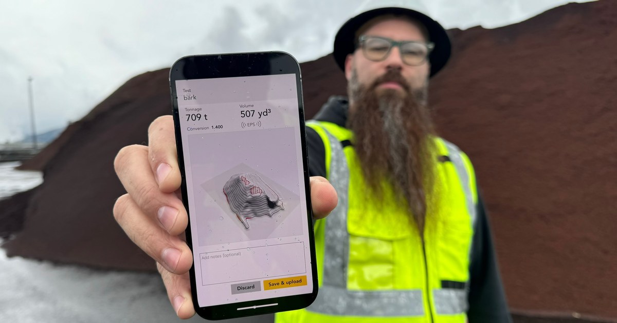 A worker in a high-visibility vest holds up a phone showing a completed measurement of 709 tons and 507 cubic yards, standing in front of the bark pile it measured.